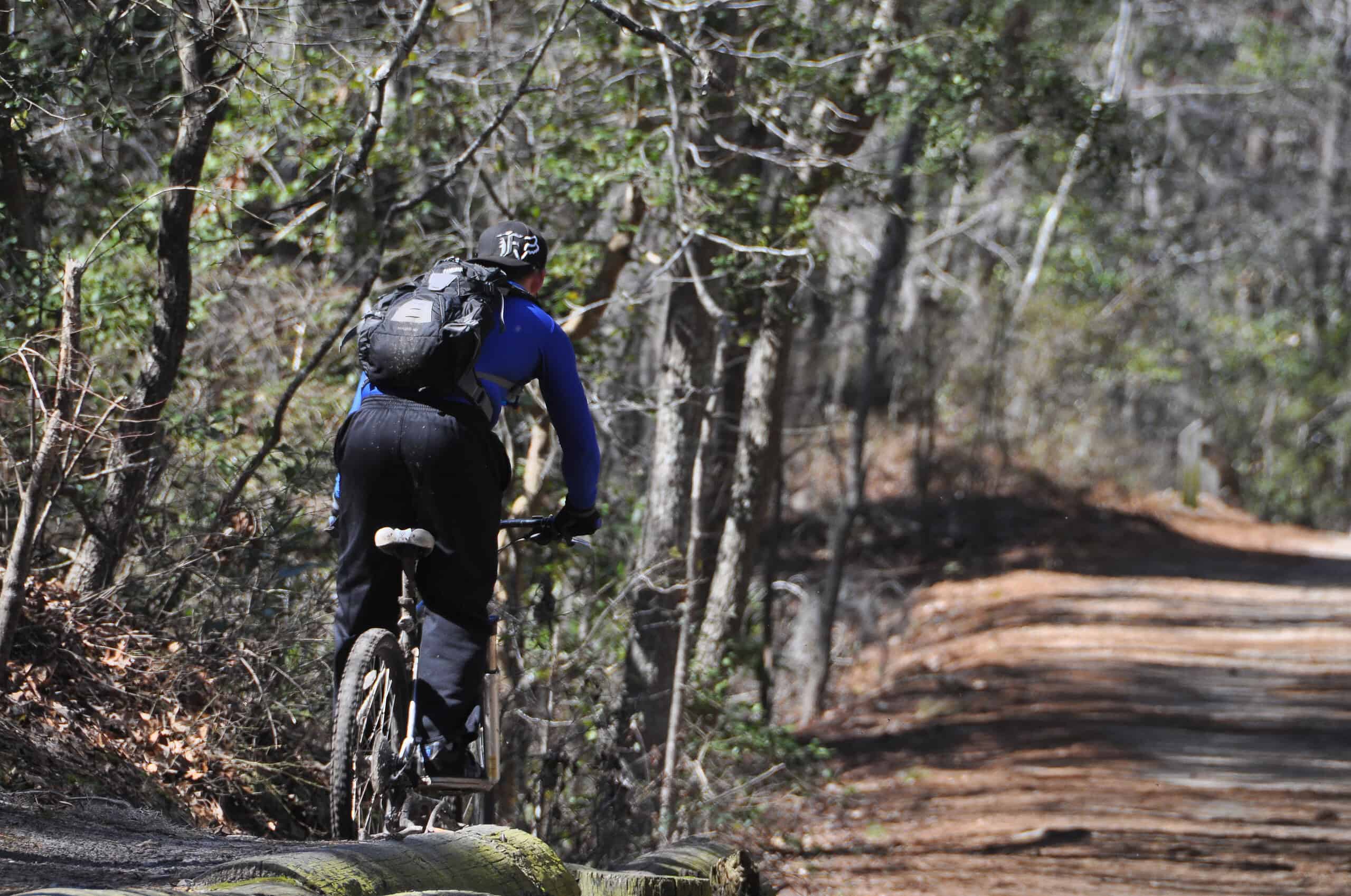 Person in blue jacket and black pants cycling through a wooded trail, carrying a backpack.