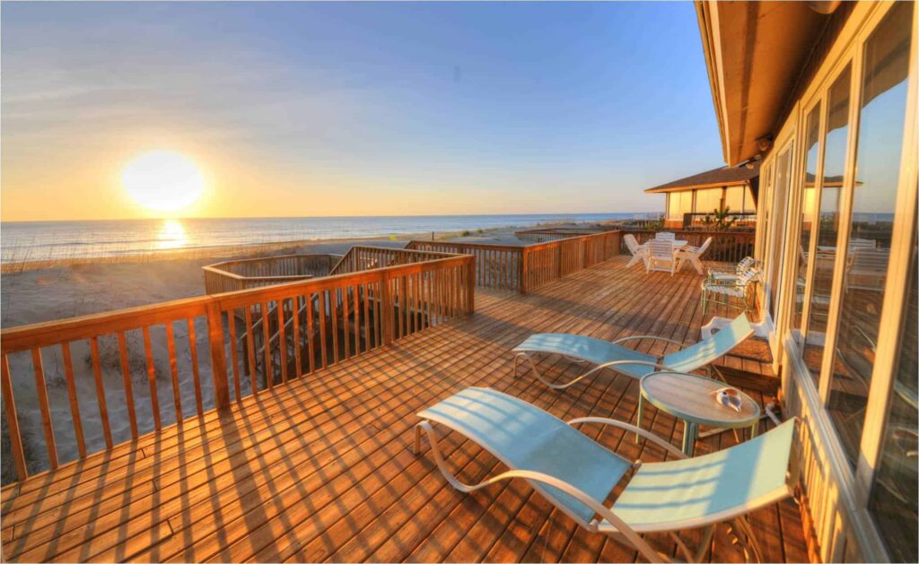 Large wooden deck with lounge chairs and tables overlooks a beach at sunset. A house with glass doors stands adjacent, offering a view of the ocean's horizon and golden sky.