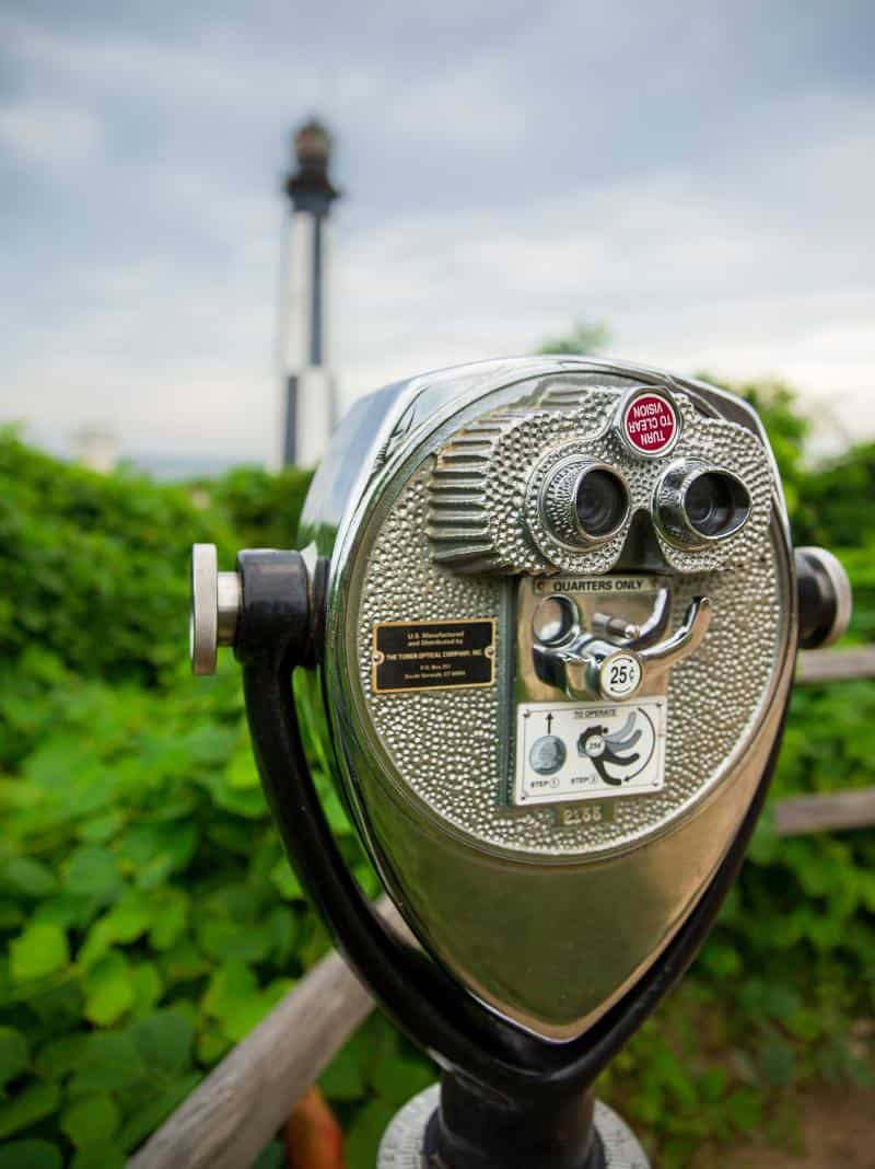Coin-operated binoculars with a lighthouse in the blurred background, surrounded by greenery and under a cloudy sky.