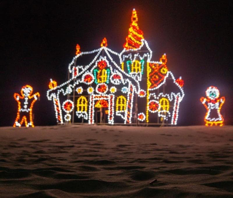 Brightly lit gingerbread house and figures made of Christmas lights on a sandy surface against a dark night sky.