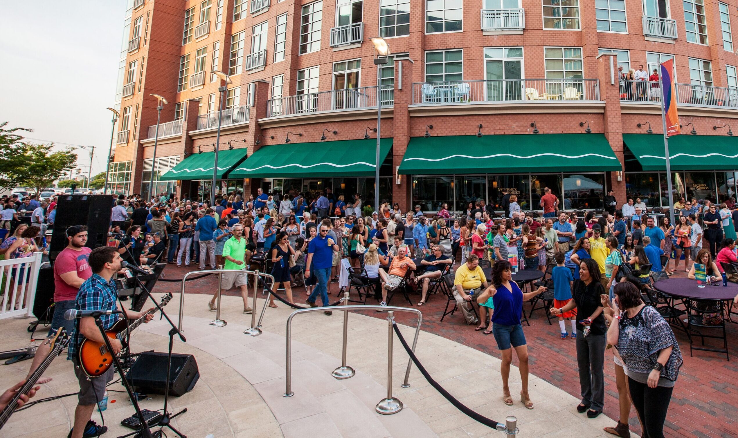 Outdoor concert with a large audience gathered in front of a brick building with green awnings. Musicians perform live music on a small stage in the foreground.