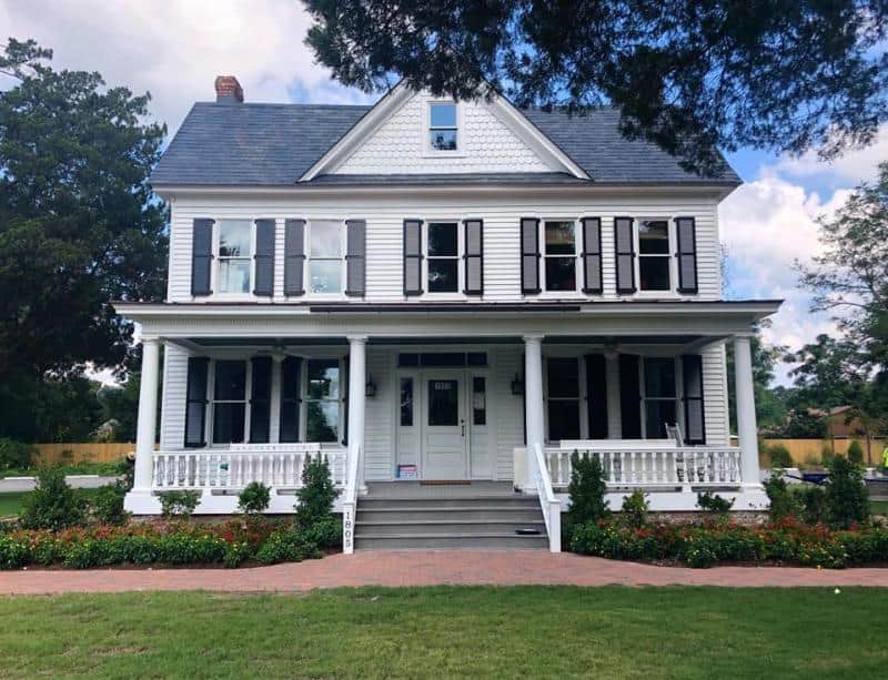 A large, two-story white house with black shutters, a covered porch, and a manicured lawn with flower beds. Trees frame the view, and a blue sky is in the background.