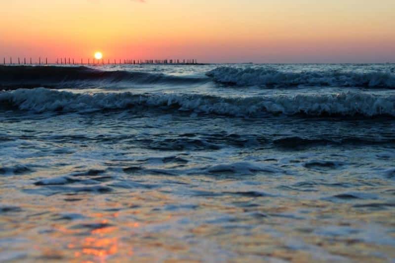 Sunset view over the ocean with waves in the foreground and a clear sky, as the sun nears the horizon.