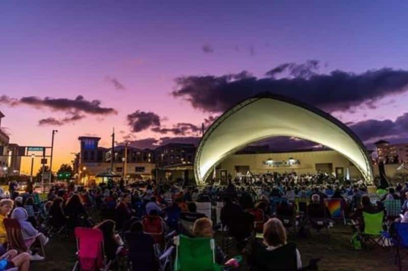 Outdoor concert at sunset with audience seated on grass in front of an illuminated, modern stage.