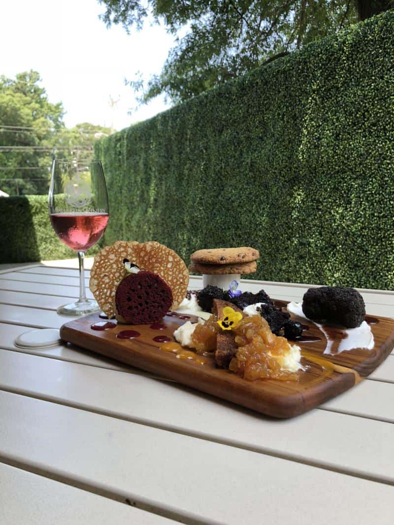 A wooden board with assorted desserts, a glass of rosé wine, and greenery in the background on a white table.