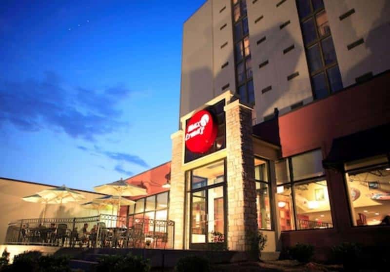 Restaurant with outdoor seating and umbrellas at dusk, featuring a red circular logo on a stone column, attached to a multi-story building.