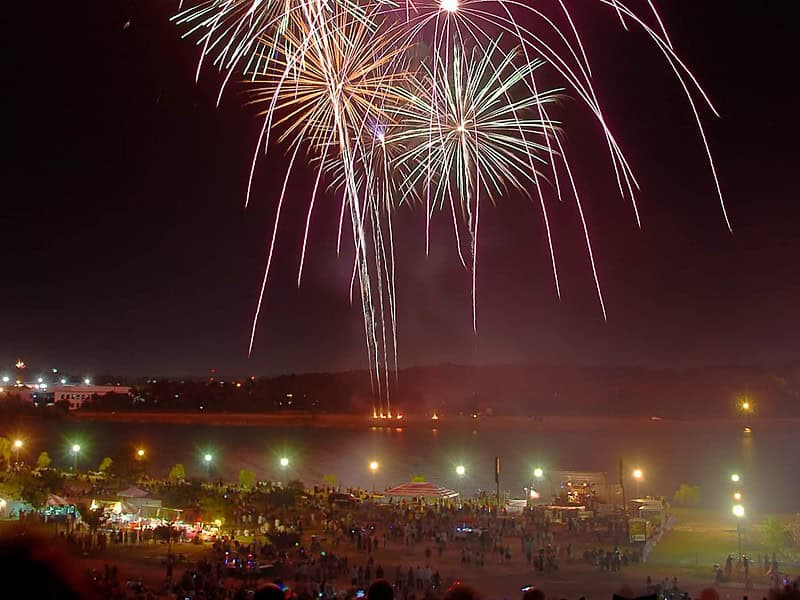 Fireworks explode in the night sky over a large crowd gathered by a riverbank, with colorful lights and stalls visible in the foreground.