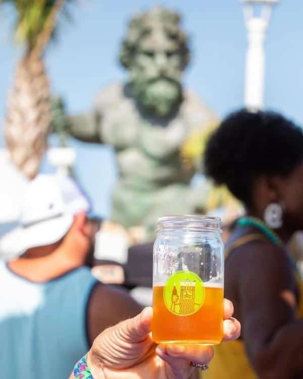 A hand holding a jar of orange beverage with a logo. In the background, people and a large statue of a muscular figure are visible. Sunlit outdoor setting.
