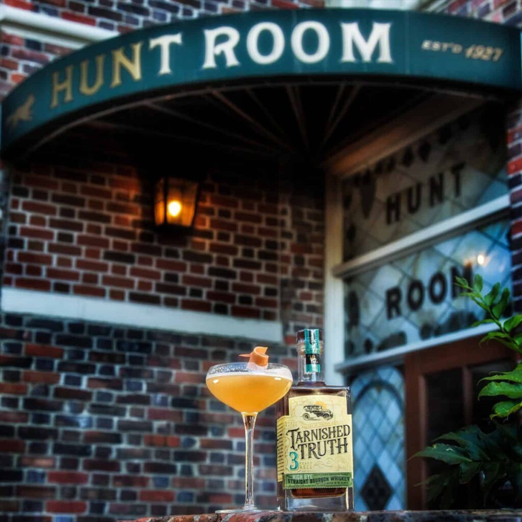 A bottle of Tarnished Truth bourbon and a cocktail glass with an orange twist sit on a ledge outside Hunt Room, a brick building with a vintage canopy.