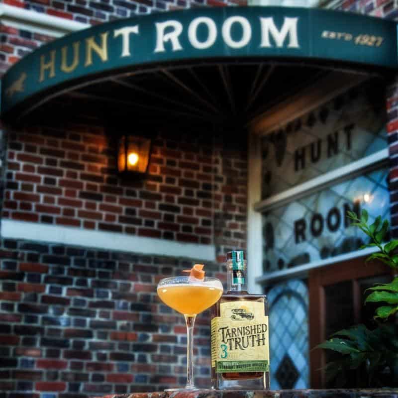 Cocktail glass and Tarnished Truth whiskey bottle in front of Hunt Room's brick exterior with signage.