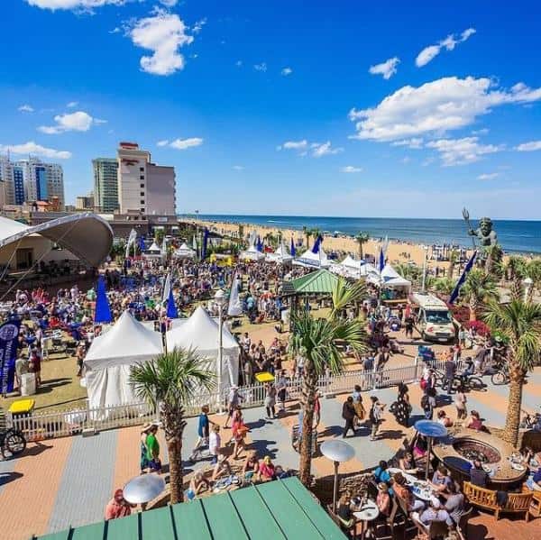 Crowded beachside festival with tents, vendors, and visitors near a sandy beach and ocean under a bright blue sky.
