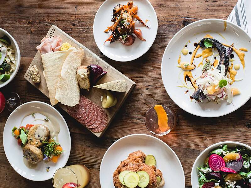 Aerial view of a wooden table with various dishes, including salads, charcuterie, bread, and drinks, arranged in a casual dining setting.