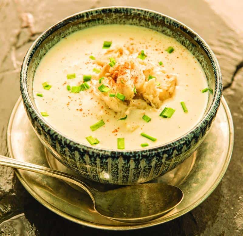 A bowl of creamy soup garnished with chives and spices, served on a saucer with a spoon beside it.