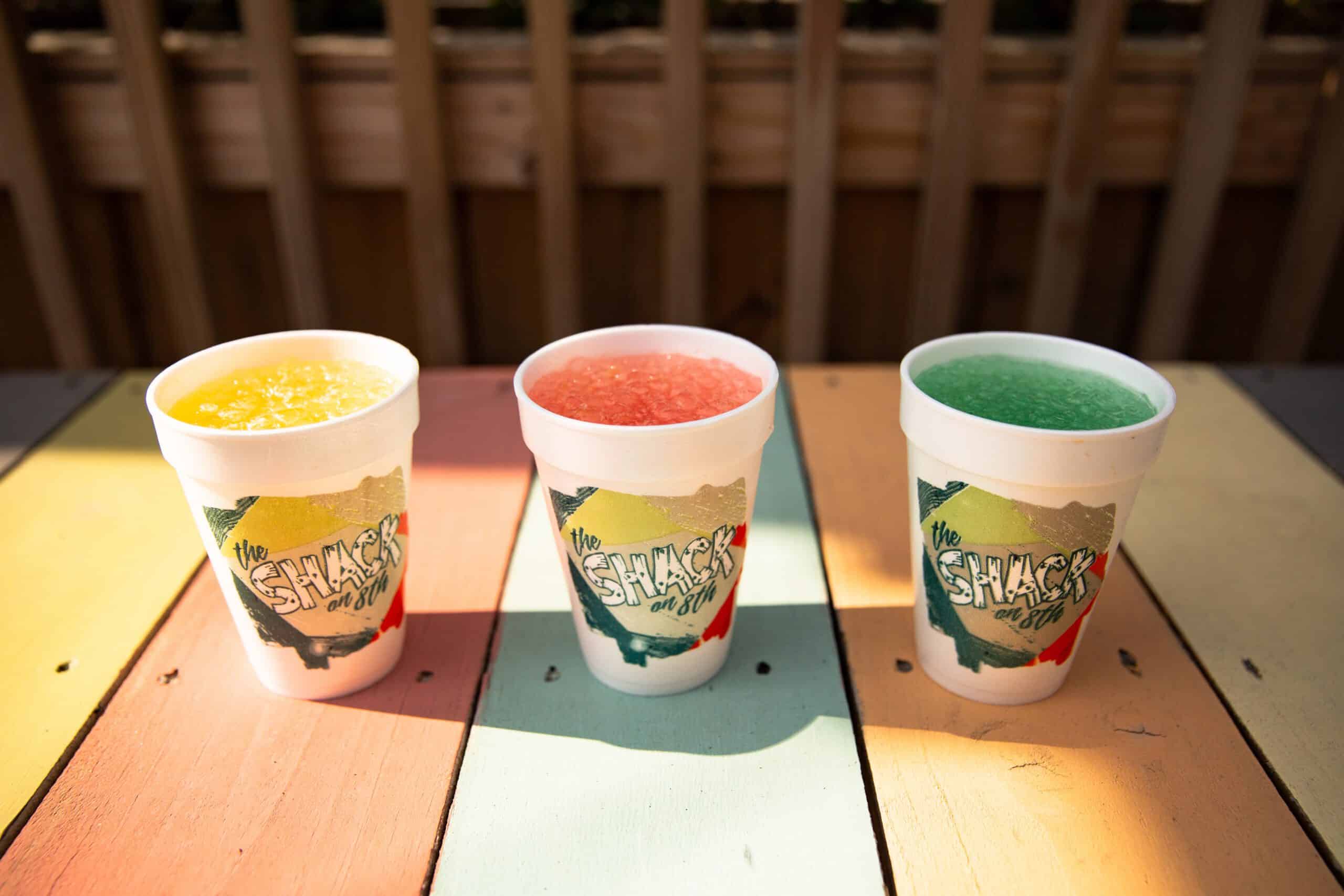 Three colorful slush drinks in white cups with "The Shack on 8th" logo, placed on a multicolored wooden table, in bright sunlight.