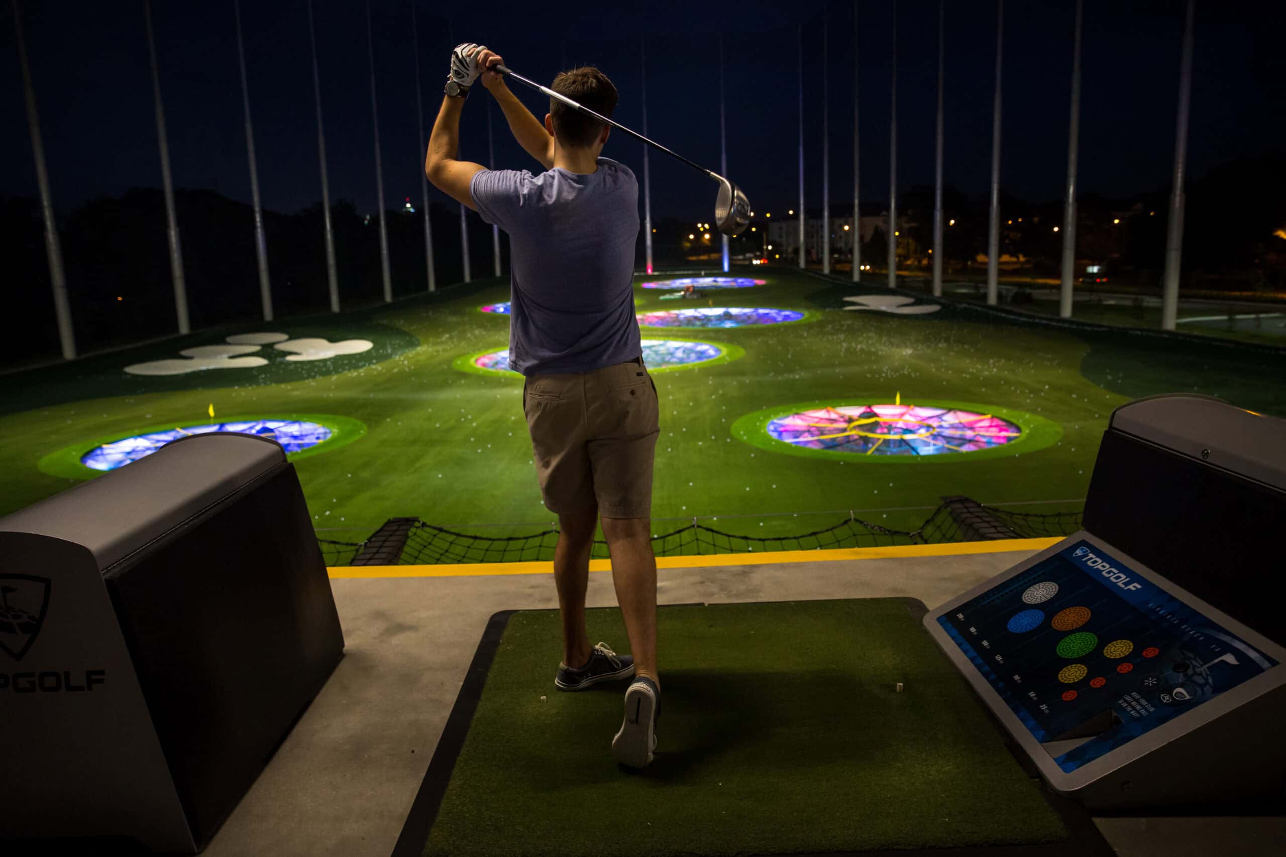 A man swings a golf club on an illuminated driving range with colorful targets at night.