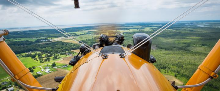 View from an open cockpit biplane flying over a rural landscape with fields and forests under a cloudy sky.