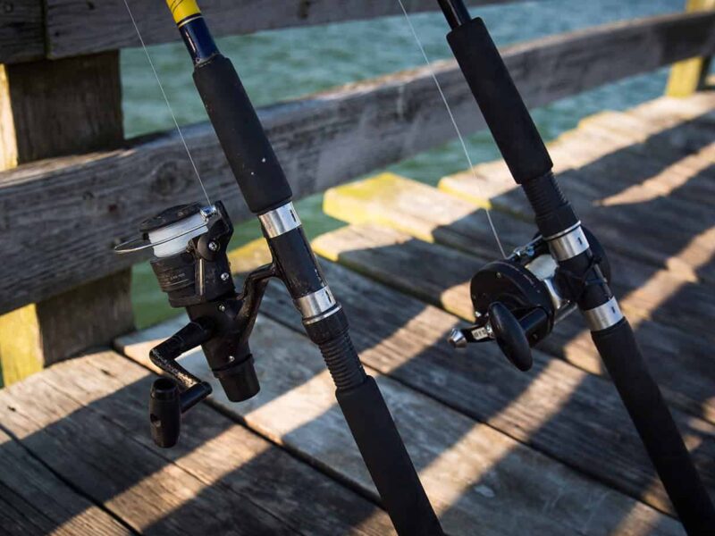 Two fishing rods leaning against a wooden railing on a sunny pier, with water visible in the background.