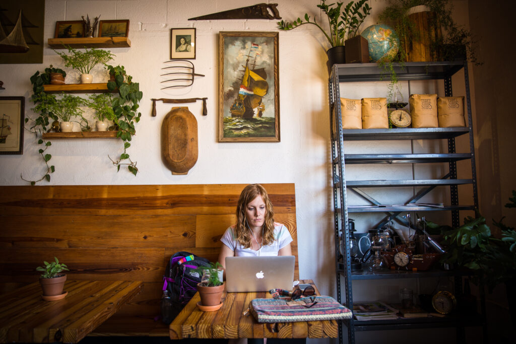 A person uses a laptop at a wooden table in a cozy cafe. The wall is decorated with nautical-themed items and plants. Nearby shelves hold bags and a globe.