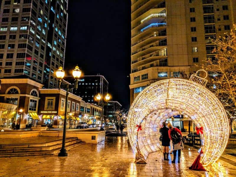 A nighttime city scene with two people walking under a large illuminated ornament decoration on a wet street, surrounded by tall buildings and lit street lamps.