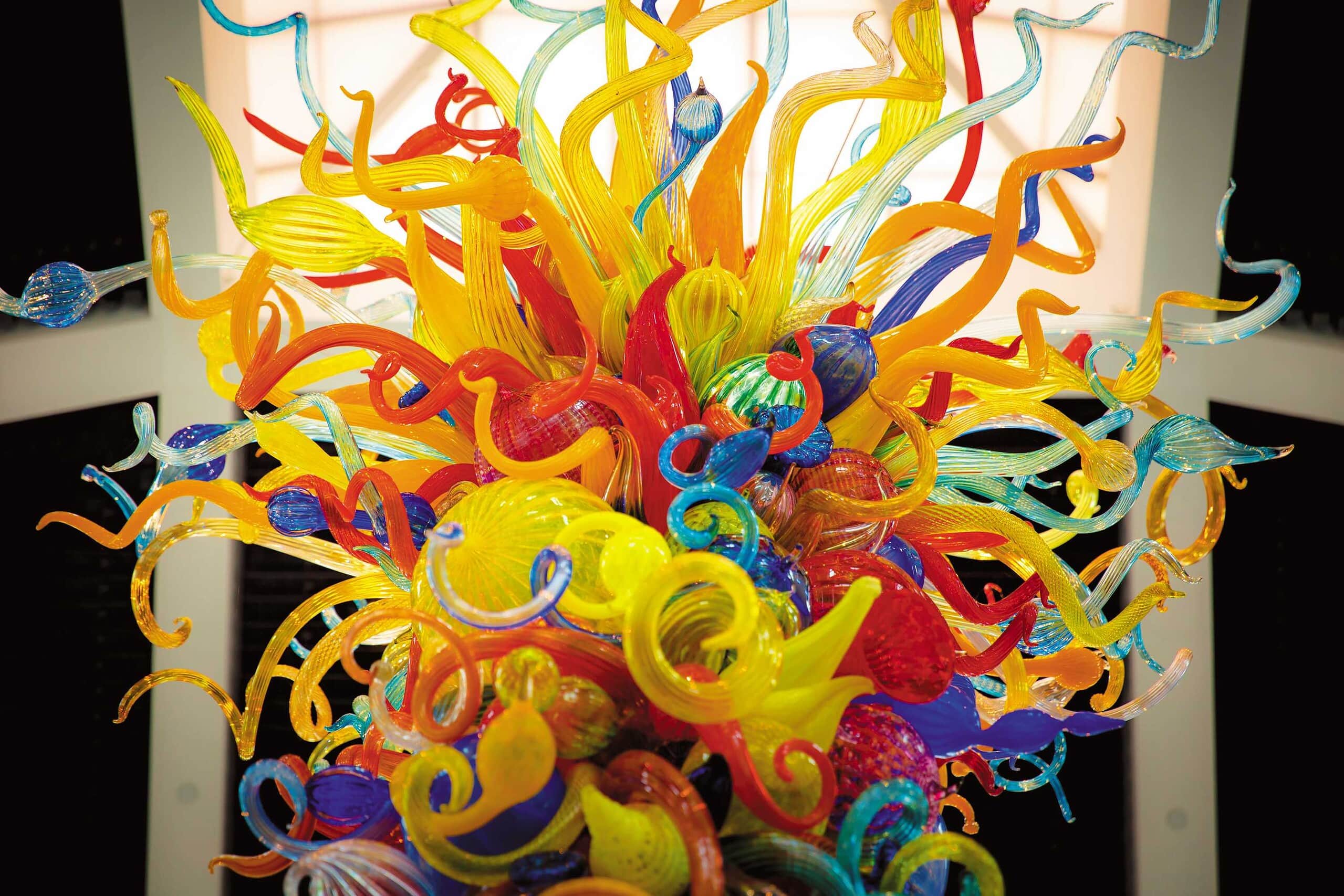 Colorful blown-glass sculpture with swirling, twisting shapes in yellow, orange, blue, green, and red, displayed under bright lighting.