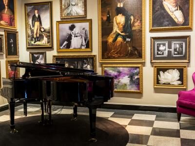 A grand piano sits in a room with checkered flooring, surrounded by numerous framed paintings and photographs on the walls. Two magenta armchairs and a small table with a lamp are nearby.