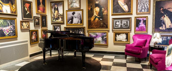 A grand piano sits in a room with checkered flooring, surrounded by numerous framed paintings and photographs on the walls. Two magenta armchairs and a small table with a lamp are nearby.