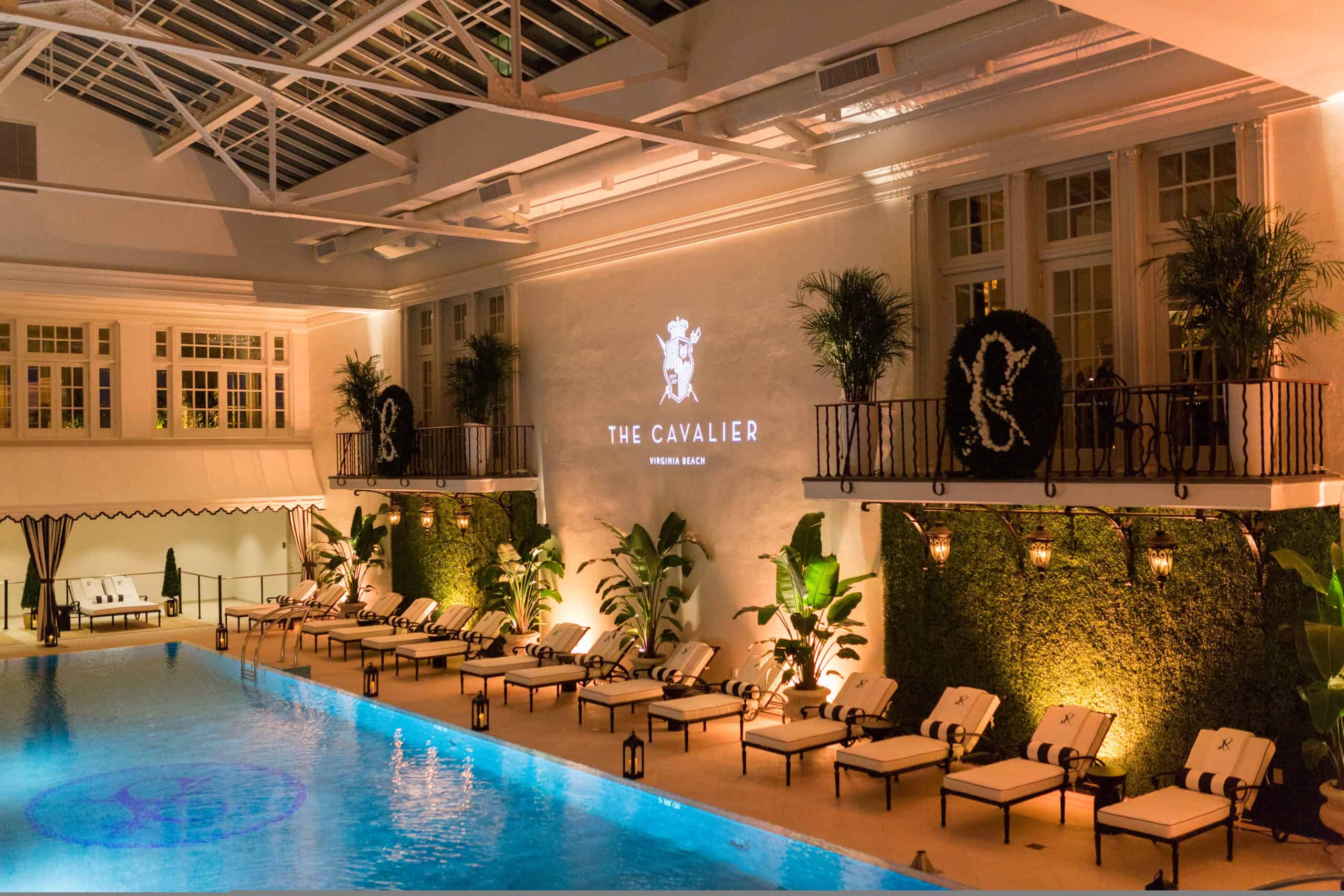 Indoor pool area with lounge chairs and greenery on both sides. "The Cavalier" logo is projected on the wall above. Ceiling features glass panels for natural light.