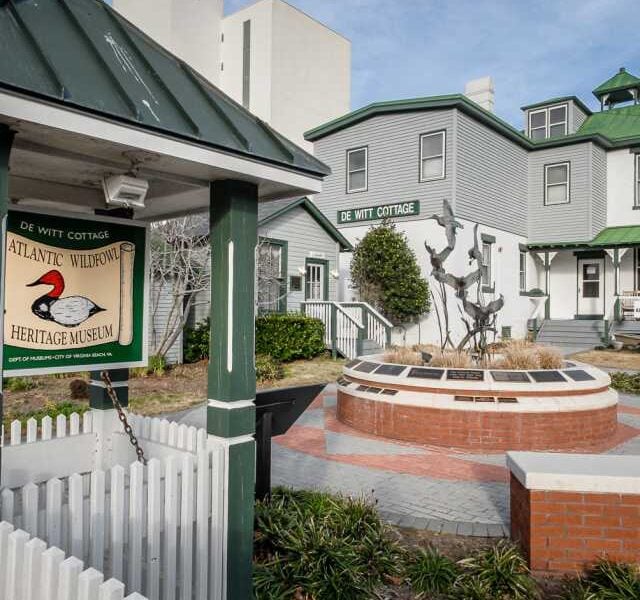 The exterior of the DeWitt Cottage and Atlantic Wildfowl Heritage Museum, with a sign, white fence, and metal sculpture in a circular brick courtyard.