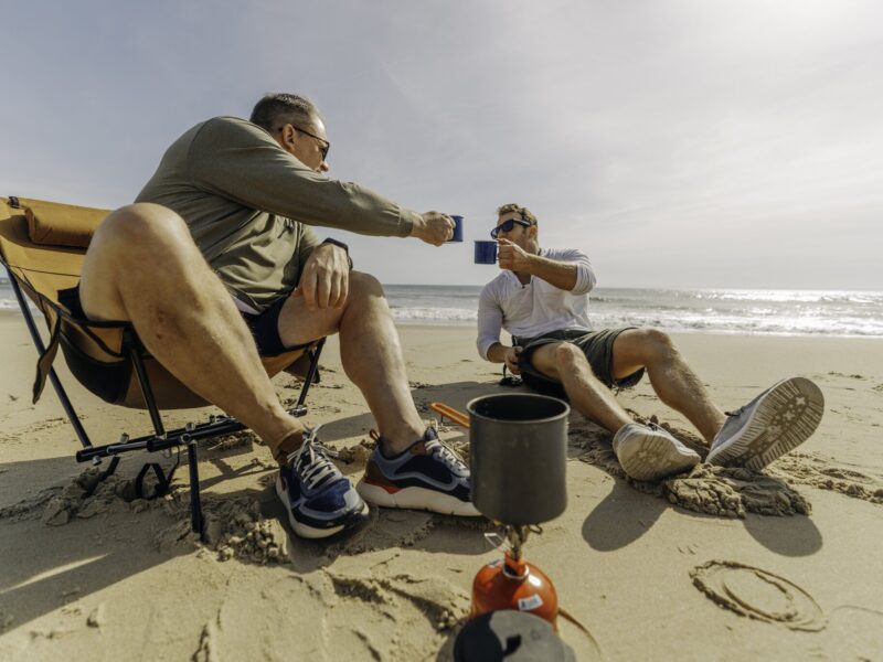 Two men enjoying drinks on the beach, seated in camping chairs with a portable stovetop beside them.