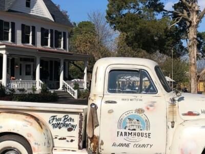 A vintage white truck with rustic branding is parked in front of a white farmhouse. Trees and a few people are in the background.