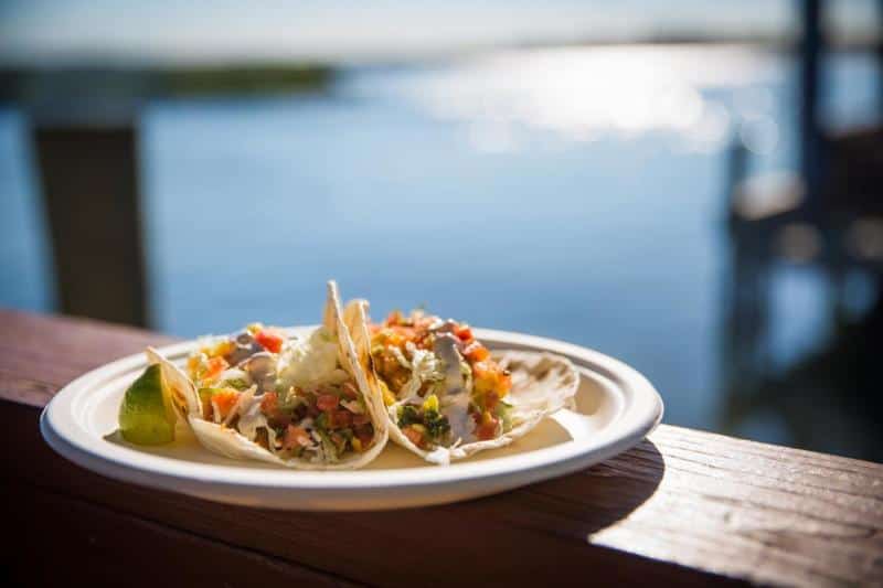 Two tacos with a lime wedge on a white plate, placed on a wooden railing overlooking a sunny waterfront.