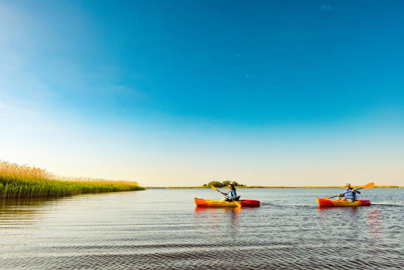 Two people kayaking on a calm lake with clear blue skies. Grassy shorelines and distant trees are visible.