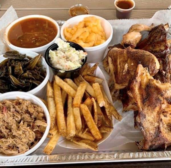A platter with grilled meats, fries, coleslaw, collard greens, mac and cheese, and soup in a cup, arranged on a silver tray.