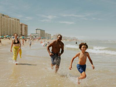 A man, woman, and boy run through shallow ocean waves on the Virginia Beach oceanfront. In the background, people relax under umbrellas near tall hotel and apartment buildings.