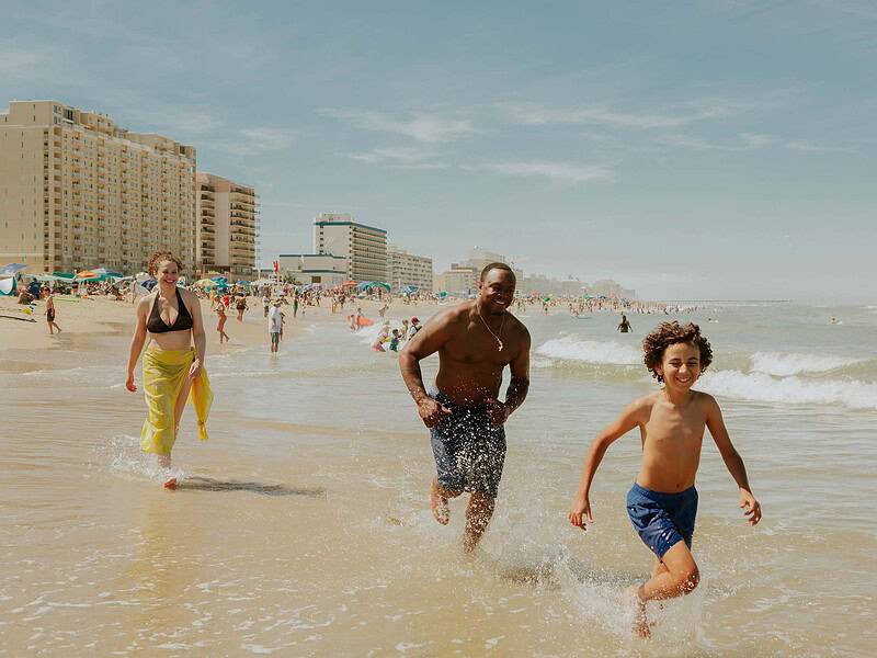 A man, woman, and boy run through shallow ocean waves on the Virginia Beach oceanfront. In the background, people relax under umbrellas near tall hotel and apartment buildings.