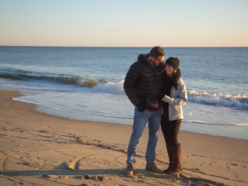 A couple stands close together on a beach at sunset, looking at each other.