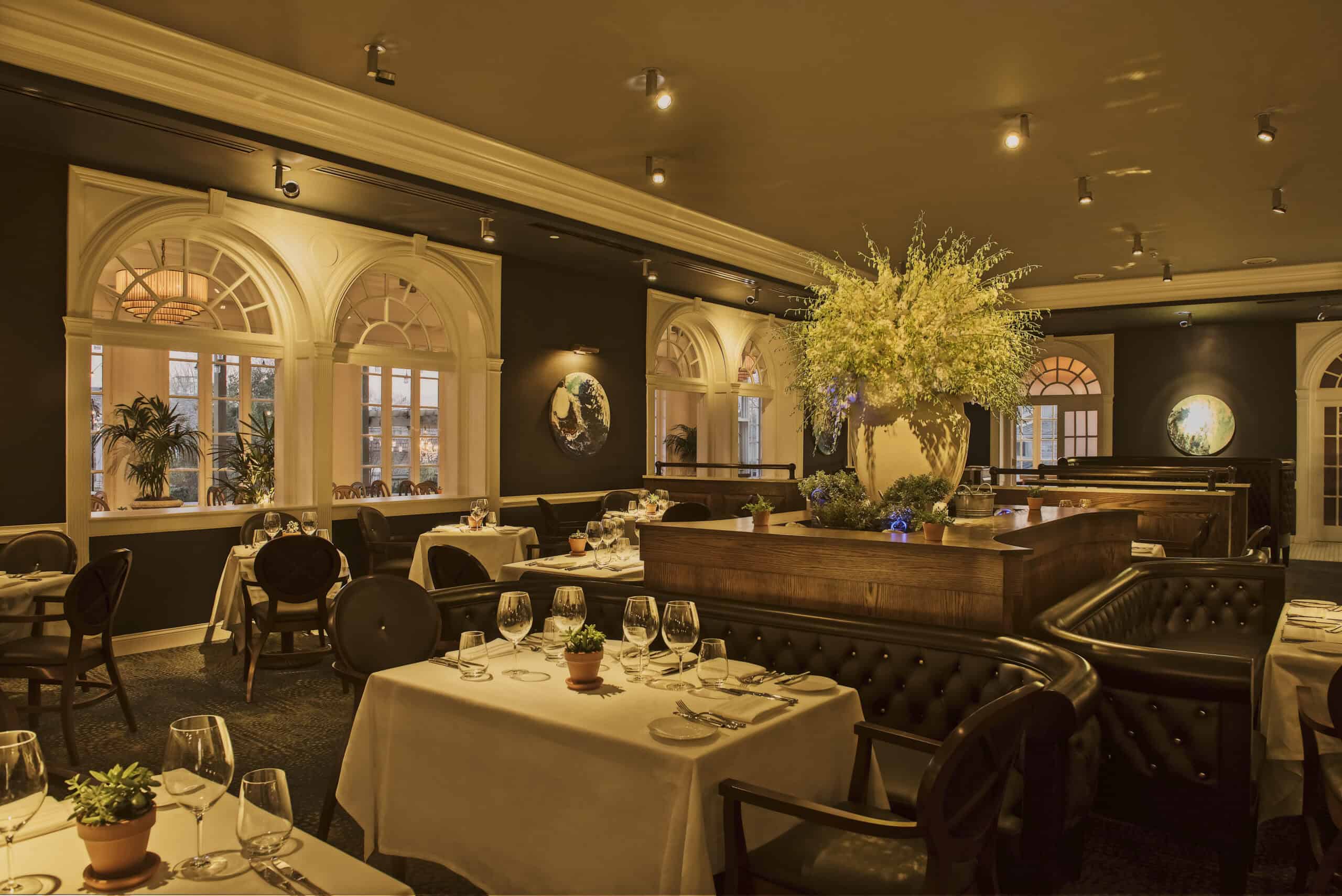 Elegant restaurant interior with white-clothed tables, cushioned chairs, and warm lighting. Large floral centerpiece on a dark wood counter. Arched windows in the background.