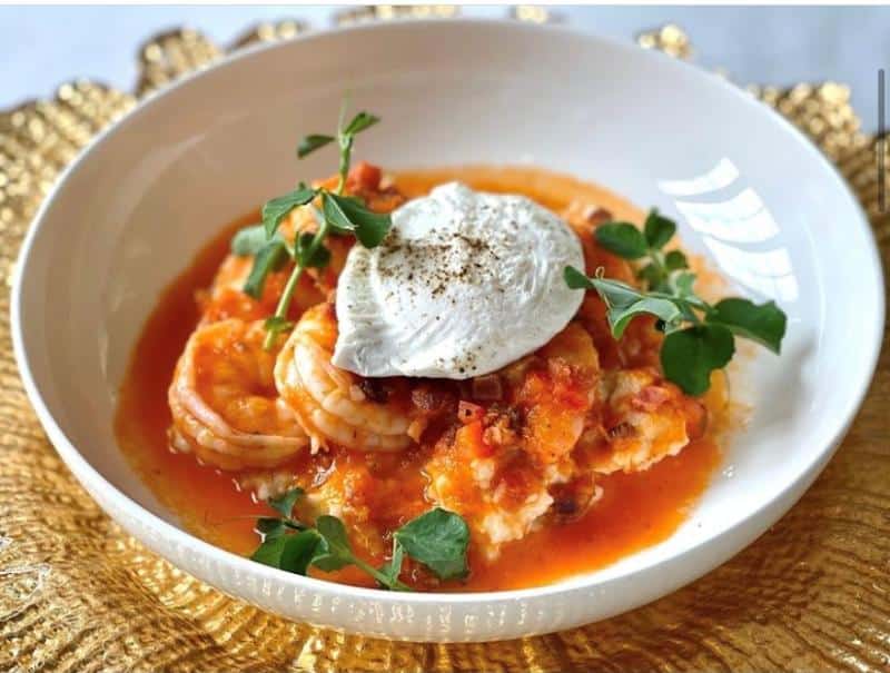A white bowl containing shrimp in red sauce, topped with a poached egg and garnished with green leaves, placed on a decorative golden surface.