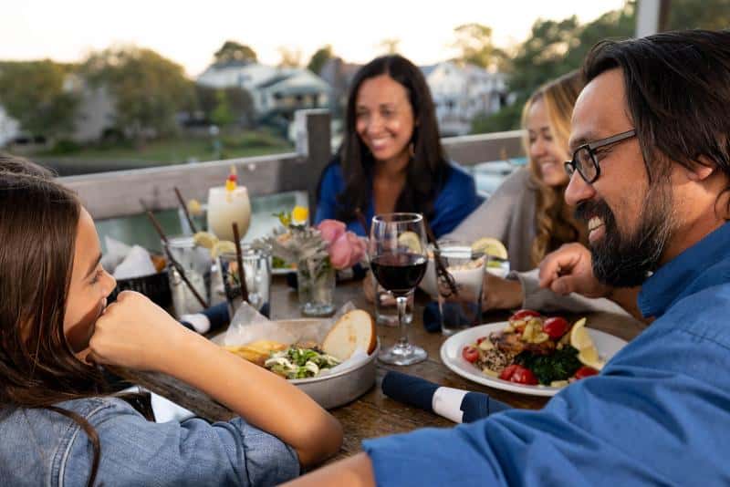 A group of people sitting at an outdoor table enjoying a meal together, with plates of food, drinks, and a scenic backdrop of trees and houses.