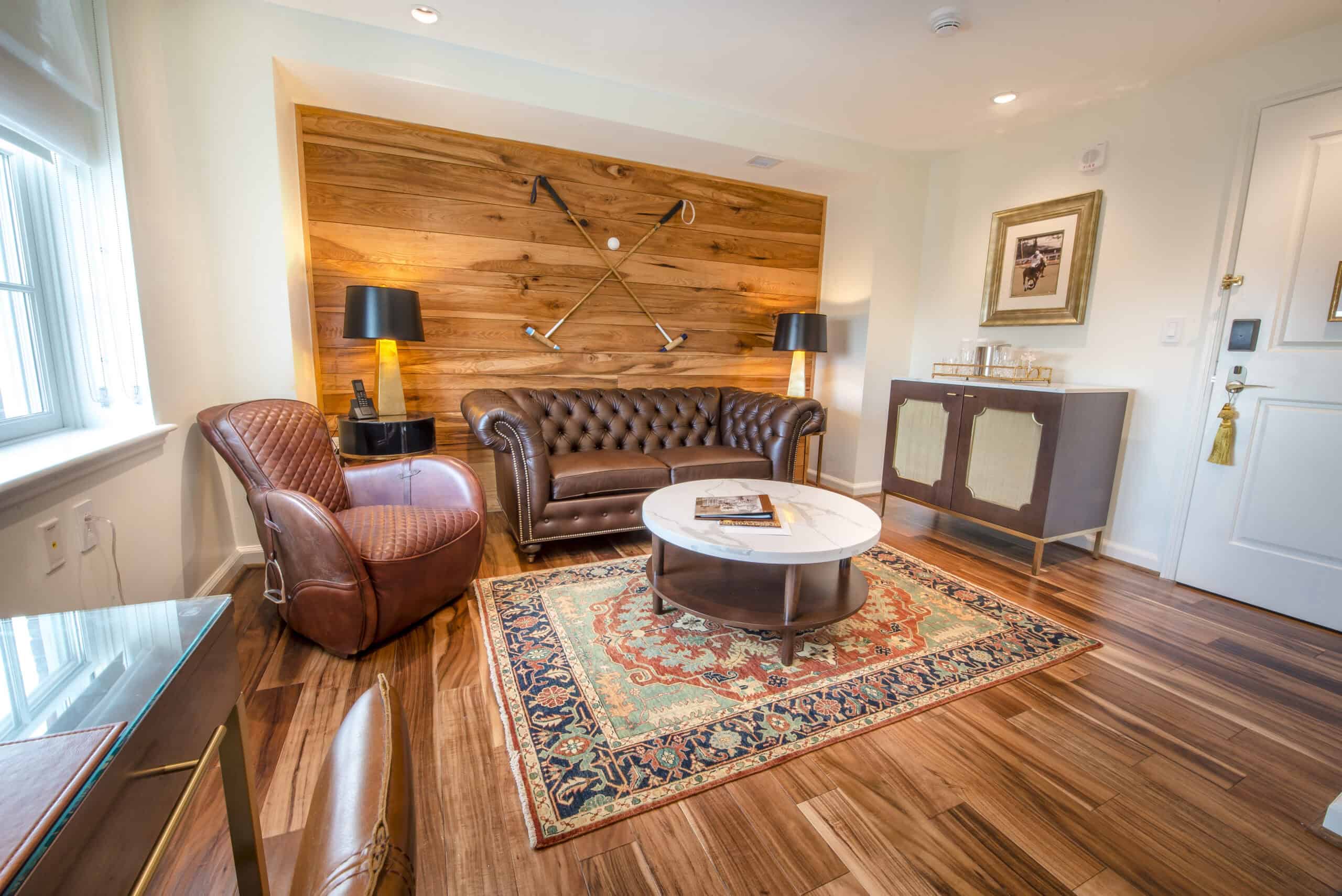 A cozy living room with a leather sofa, armchair, and round coffee table on a patterned rug. Golf clubs decor on a wooden accent wall. Floor lamp and sideboard with framed picture.