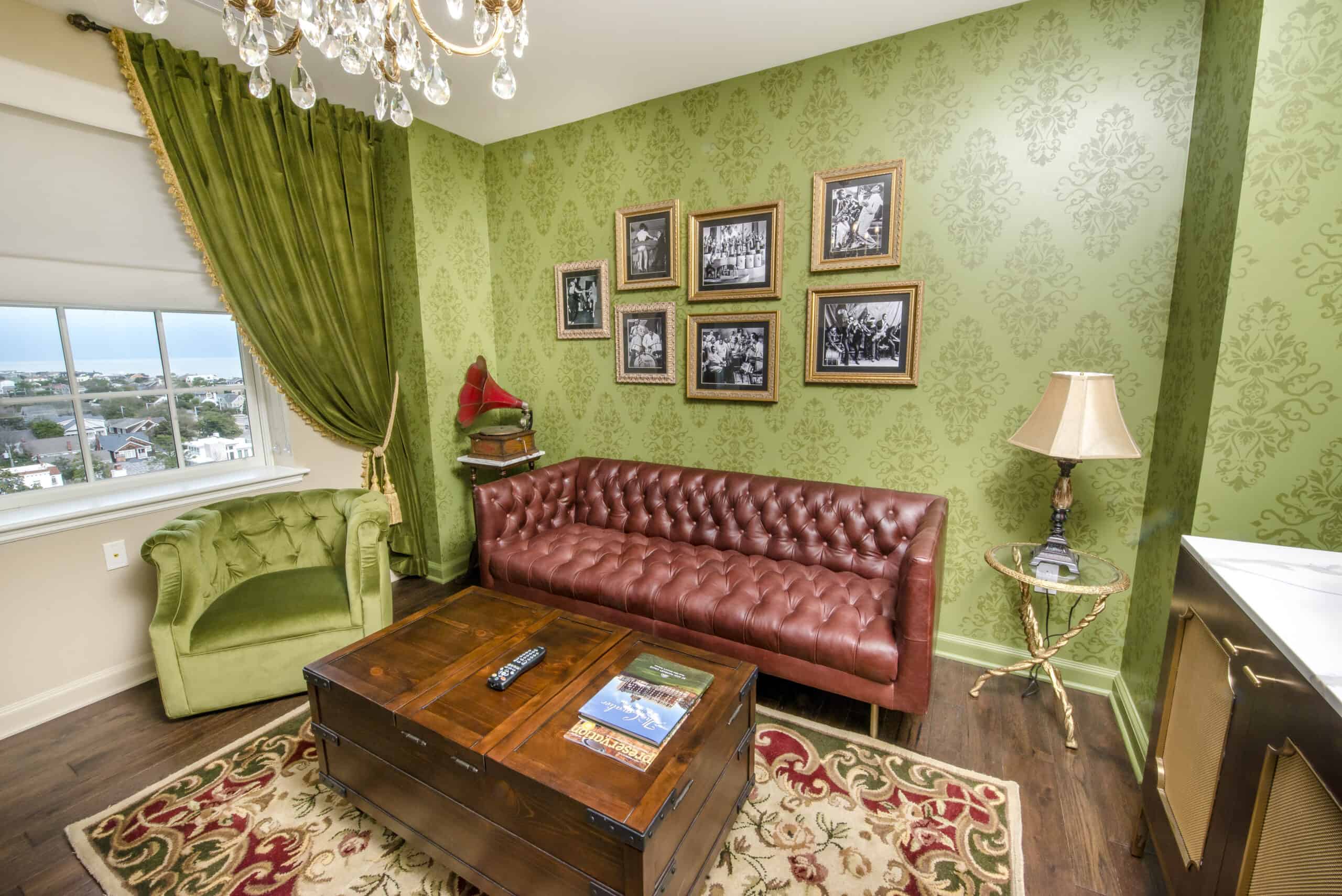 Room with green damask wallpaper, brown leather sofa, green armchair, wooden coffee table, and multiple framed pictures on the wall. A chandelier and lamp provide lighting.