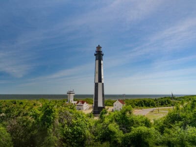 Cape Henry Lighthouse: A tall black and white lighthouse stands near a coast, surrounded by lush greenery and small buildings under a clear blue sky.