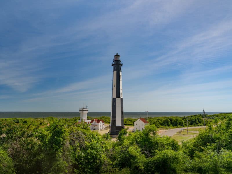 Cape Henry Lighthouse: A tall black and white lighthouse stands near a coast, surrounded by lush greenery and small buildings under a clear blue sky.