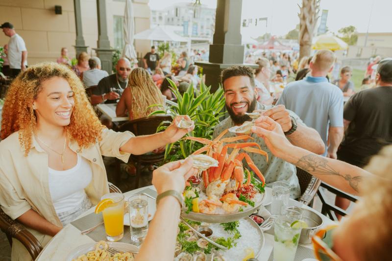 Group of people sharing a seafood platter and drinks at an outdoor restaurant with others in the background.