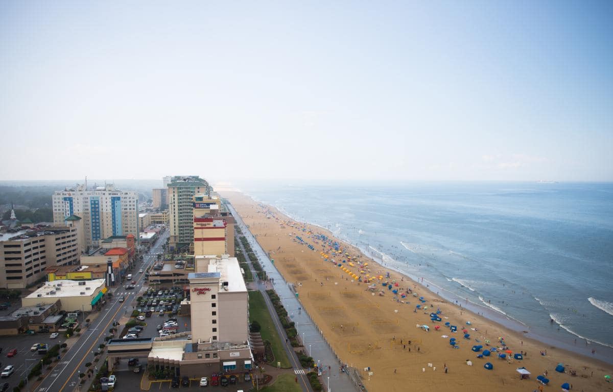 Aerial view of a beachfront cityscape with hotels on the left and a sandy beach with colorful umbrellas and people on the right, adjacent to a calm ocean.