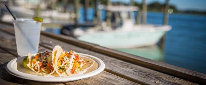 Tacos on a plate and a white drink with a straw sit on a wooden table by a docked boat and blue water.