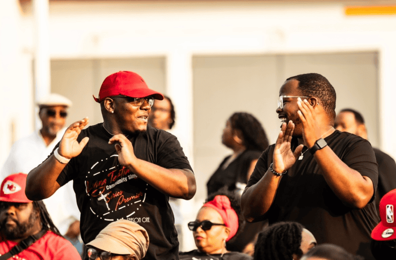 Two men stand and clap, wearing casual clothing, in an outdoor crowd. One wears a red cap and black shirt, while the other has glasses and a black shirt. People in background appear engaged.