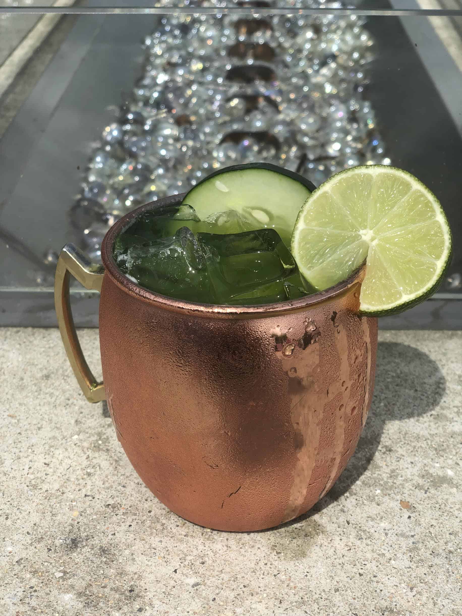 Copper mug with iced green drink, garnished with cucumber slices and a lime wedge, placed on a stone surface.
