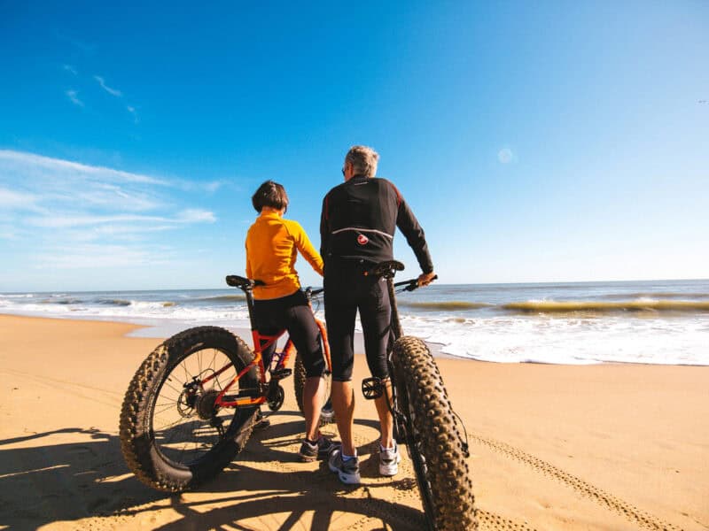 A man and woman admire the ocean while sitting on fat bikes on a sandy beach under a clear blue sky.