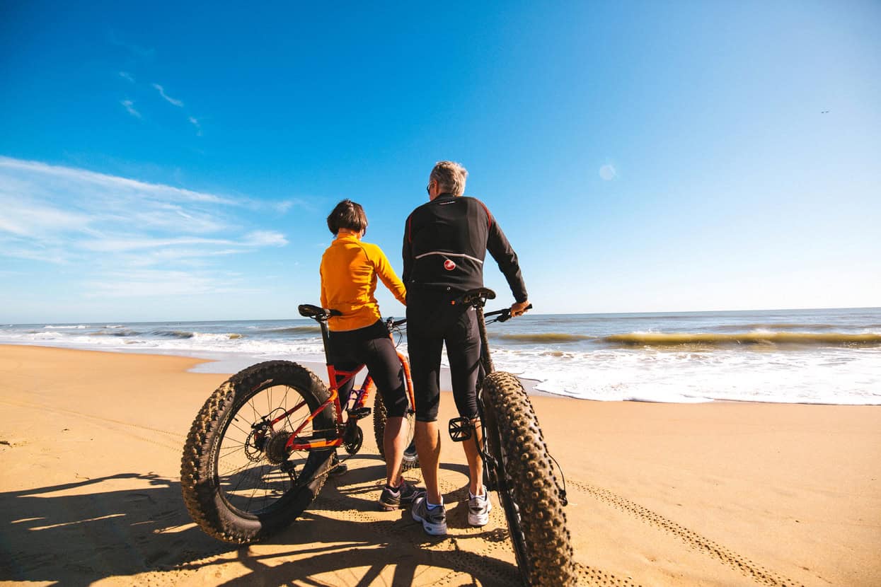 A man and woman admire the ocean while sitting on fat bikes on a sandy beach under a clear blue sky.